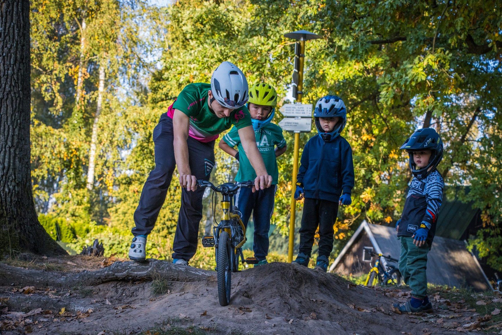 Ondřej Vít ukazuje dětem přejezd terénní vlny na dětském kole, tři děti v přilbách přihlížejí