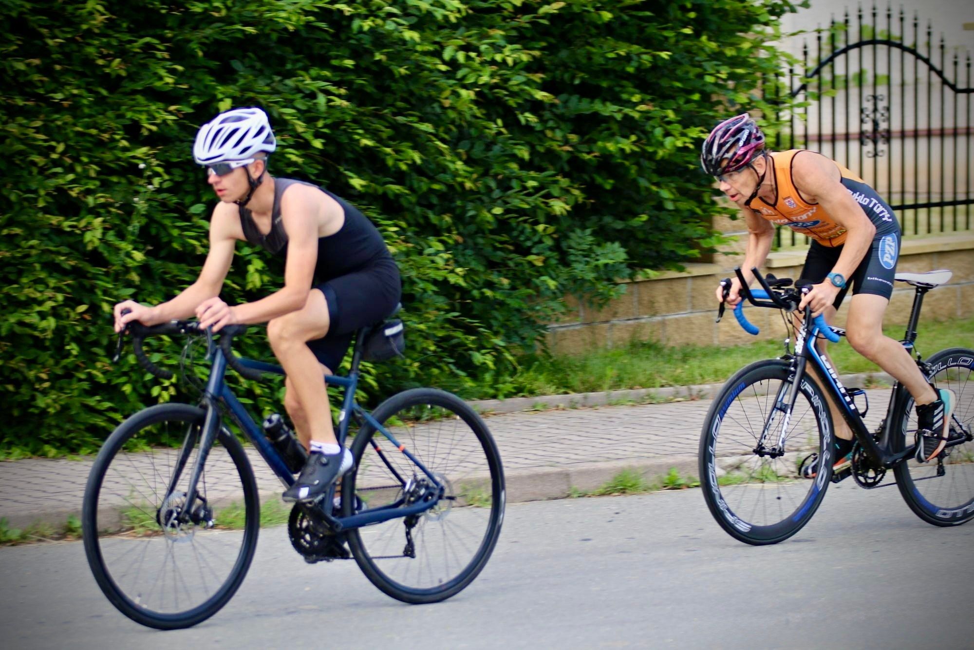 Two riders on road bikes, a young racer in a black tri suit and white helmet in front