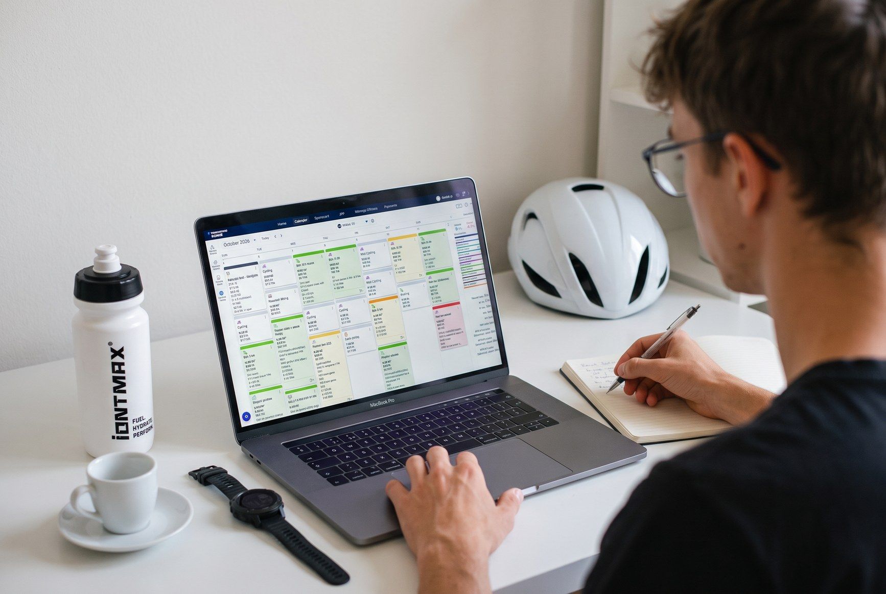 Trenér u notebooku s tréninkovým kalendářem, na stole cyklistická přilba, hodinky a láhev